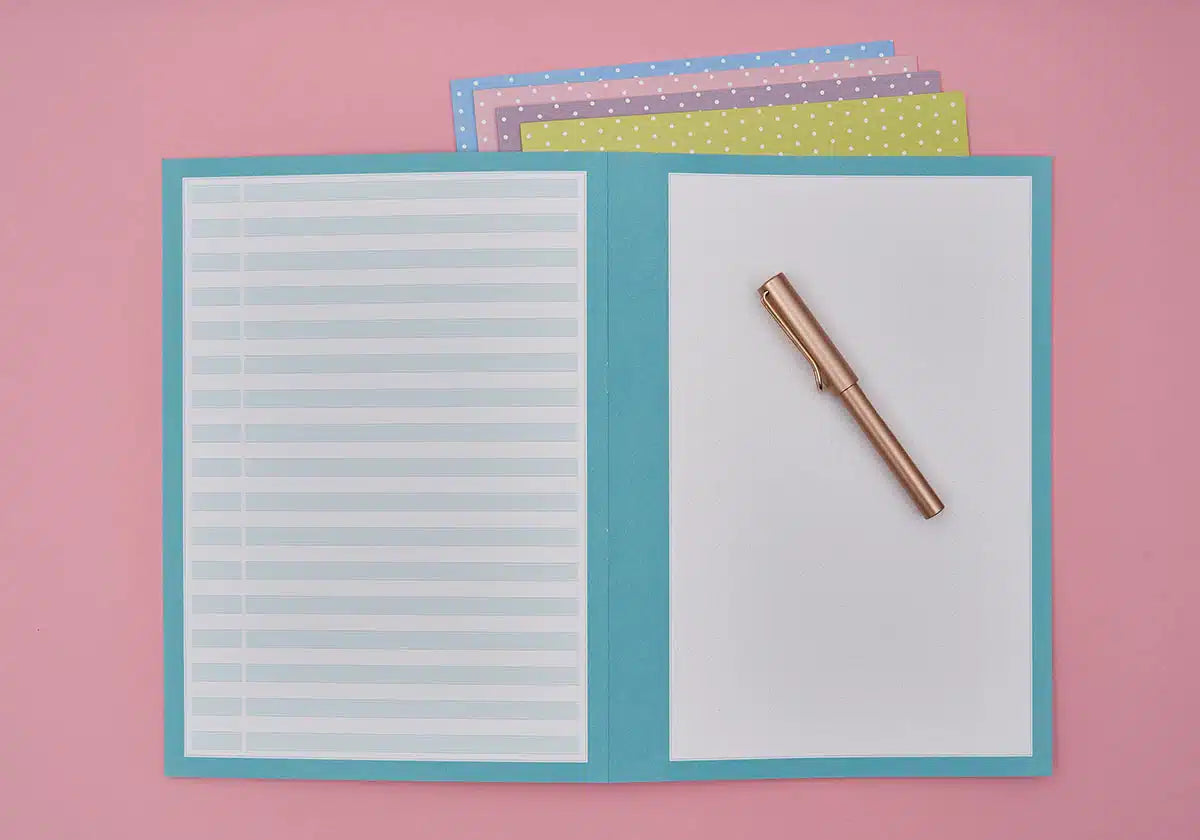 Ein türkisfarbenes Objekt aus dem amoedo Wochenmappen »Jeder Tag zählt« 5er Set zum Abheften liegt auf einer rosa Oberfläche und hält links liniertes Papier und rechts leere Blätter mit einem goldenen Stift bereit. Unter den Mappen sorgen bunt gepunktete Papiere für eine verspielte Note.
