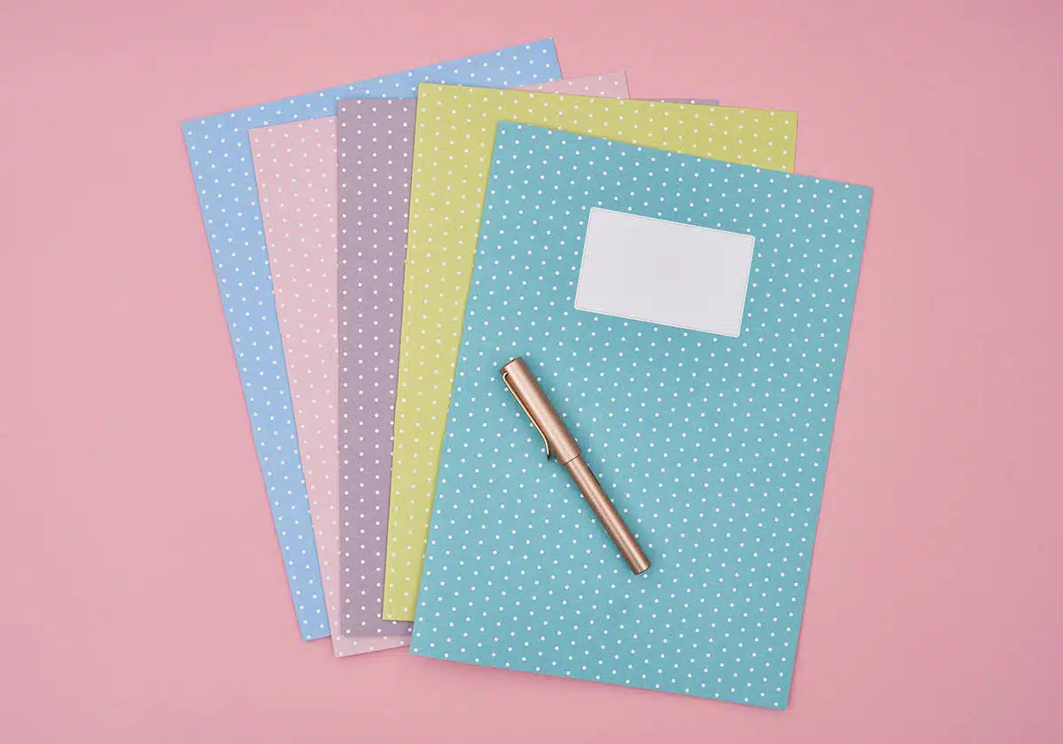 Ein Set aus fünf Notizbüchern der Wochenmappen »Jeder Tag zählt« von amoedo mit Tupfen in Pastelltönen von Blau, Rosa, Grau, Gelb und Blaugrün, die auf einer rosa Oberfläche angeordnet sind. Ein goldener Stift liegt auf dem blaugrünen Notizbuch aus dem Set, das ein leeres weißes Etikett trägt.