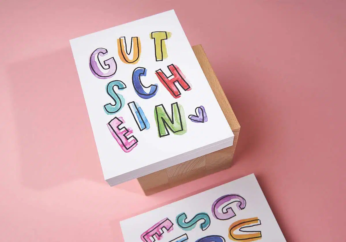 Ein Stapel Karten auf einem Holzblock, auf denen in bunten, fetten Buchstaben auf rosa Hintergrund das Wort „GUTSCHEIN“ steht. Entdecken Sie weitere einzigartige Designs aus der Postkarten-Kollektion »GUTSCHEIN« von brushmeetspaper x amoedo auf brushmeetspaper.com.