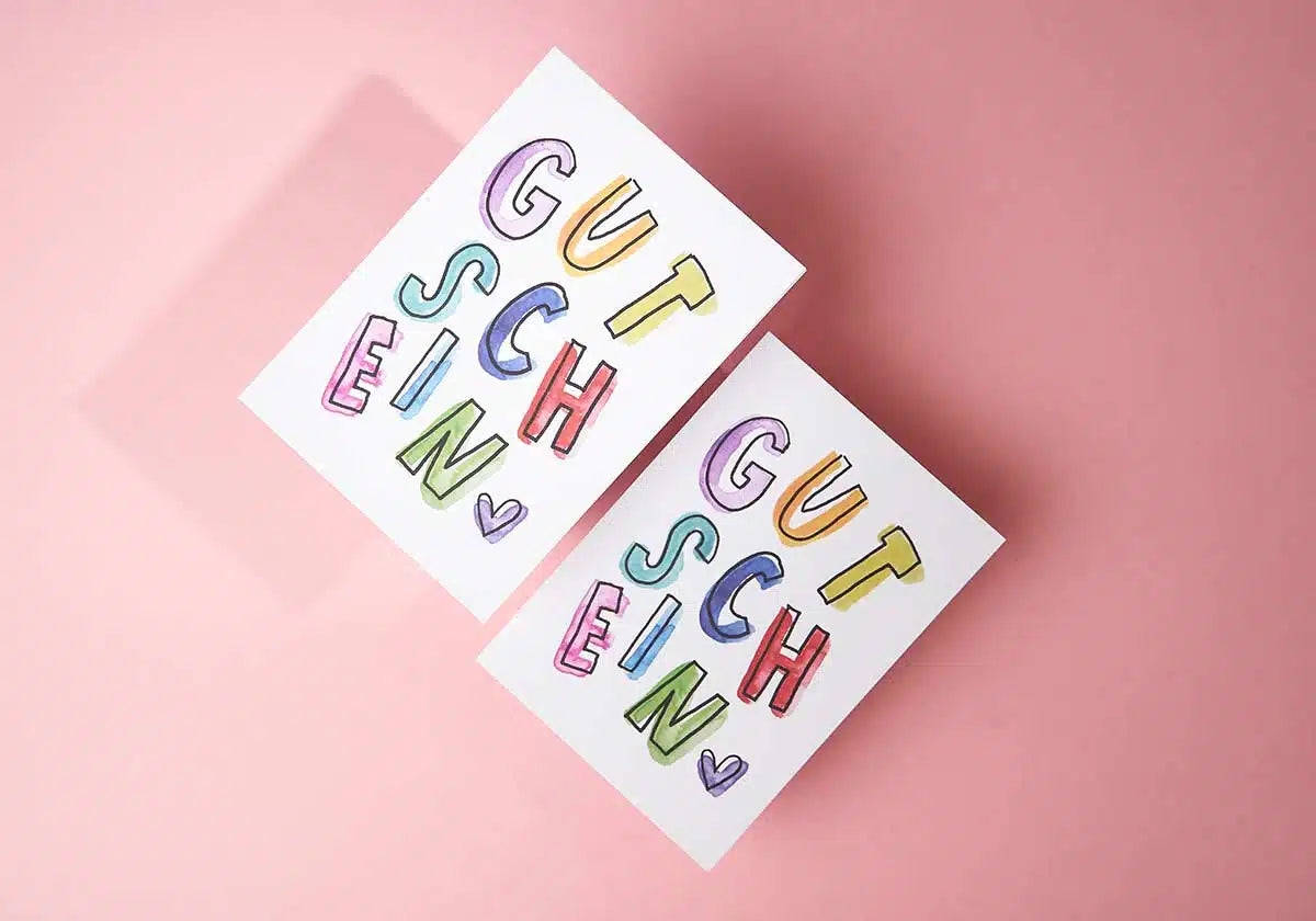 Die *Postkarte »GUTSCHEIN«* von *brushmeetspaper x amoedo* sind zwei rechteckige Karten mit verspieltem Design. Sie zeigen einen lebendigen „Gutschein“-Text und kleine Herzsymbole auf einem zartrosa Hintergrund. Der Schriftzug ist in einer skurrilen Schriftart gehalten, bei der jeder Buchstabe eine andere leuchtende Farbe hat, was den entzückenden Stil von brushmeetspaper.com widerspiegelt.