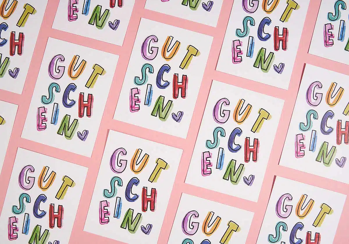 Eine lebendige Darstellung farbenfroher Postkarten auf rosa Hintergrund, jede davon zeigt das Wort „GUTSCHEIN“ in verspielten, bunten Buchstaben mit einem kleinen violetten Herz darunter, was an die skurrilen Designs von brushmeetspaper x amoedo erinnert.