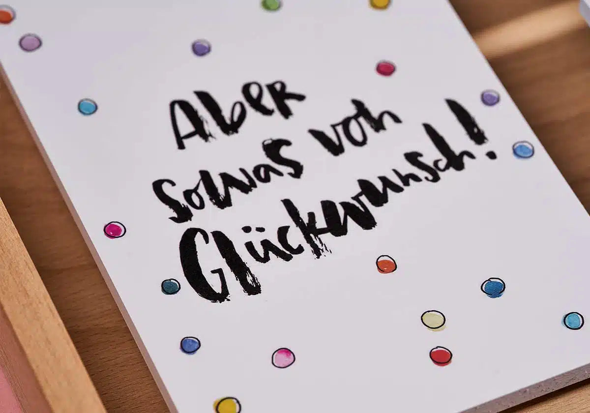 Die Postkarte »Aber sowas von Glückwunsch« vonbrushmeetspaper x amoedo zeichnet sich durch mehrfarbige Tupfen und einen handgeschriebenen deutschen Text mit der Aufschrift „Aber sowas von Glückwunsch!“ aus, der freudige Momente perfekt einfängt. Es wird elegant auf einer Holzoberfläche präsentiert.