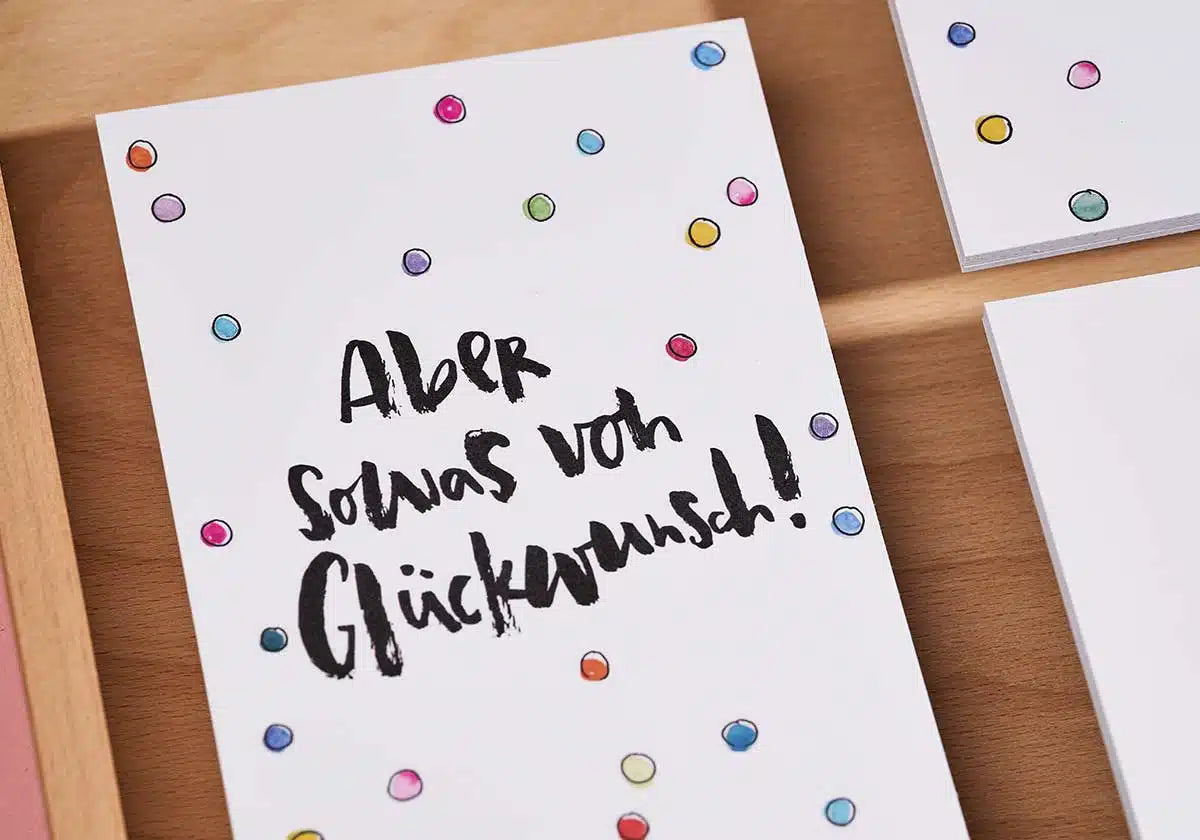 Die Postkarte „Aber sowas von Glückwunsch“ vonbrushmeetspaper x amoedo mit leuchtenden Punkten und handgeschriebenem deutschen Text fängt die Essenz freudiger Momente wunderbar ein, wenn sie auf einer Holzoberfläche ruht.