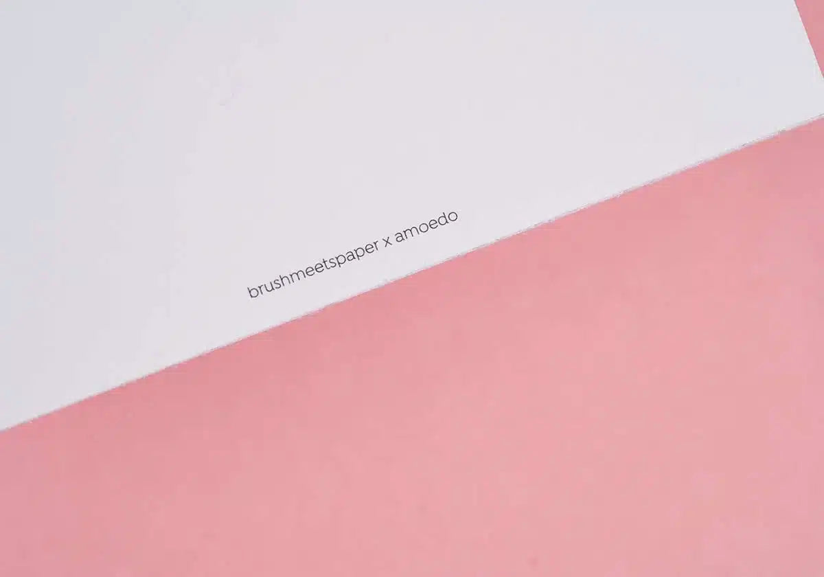Ein schräg gestelltes Blatt Papier vor rosa Hintergrund gleicht der bezaubernden Postkarte „Aber sowas von Glückwunsch“. Der Text „brushmeetspaper x amoedo“ ist anmutig am Rand aufgedruckt und fängt freudige Momente ein.