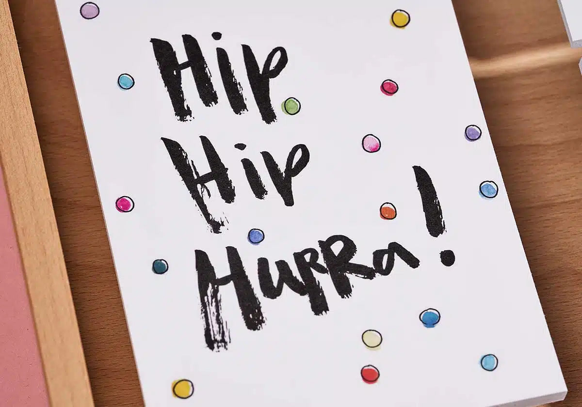 Die Postkarte »Hip Hip Hurra« von brushmeetspaper x amoedo zeigt kräftige schwarze Pinselstriche, die „Hip Hip Hurra!“ buchstabieren, und strahlt Lebendigkeit mit bunten Punkten in Blau, Rot, Orange, Grün und Lila auf einem strahlend weißen Hintergrund aus.