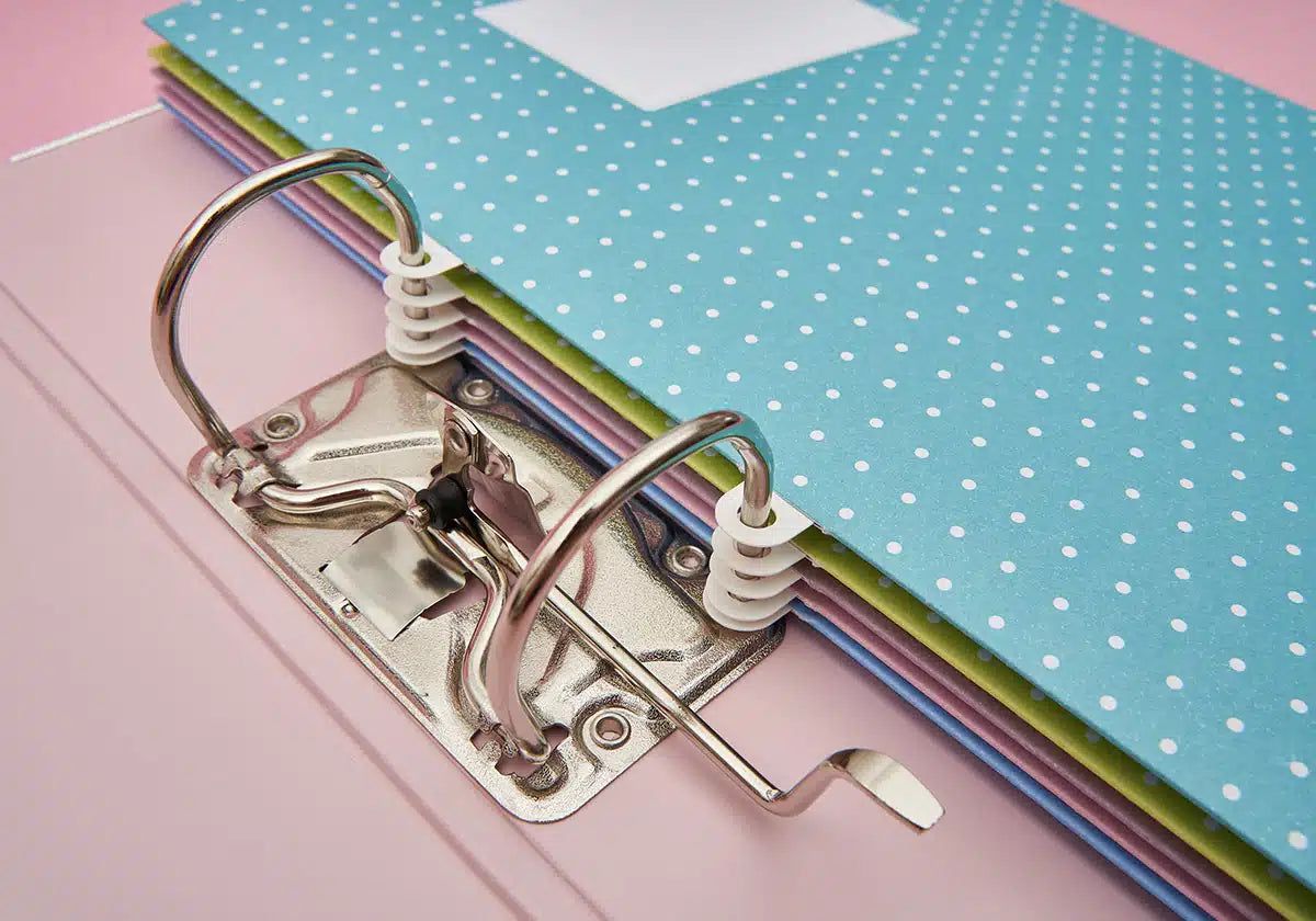 Eine Nahaufnahme eines geöffneten amoedo Wochenmappen »Jeder Tag zählt« 5er-Sets zum Abheften mit zwei Ringen auf der linken Seite, das einen Stapel bunter, gepunkteter Papiere, überwiegend blau, auf einer rosa Oberfläche hält.