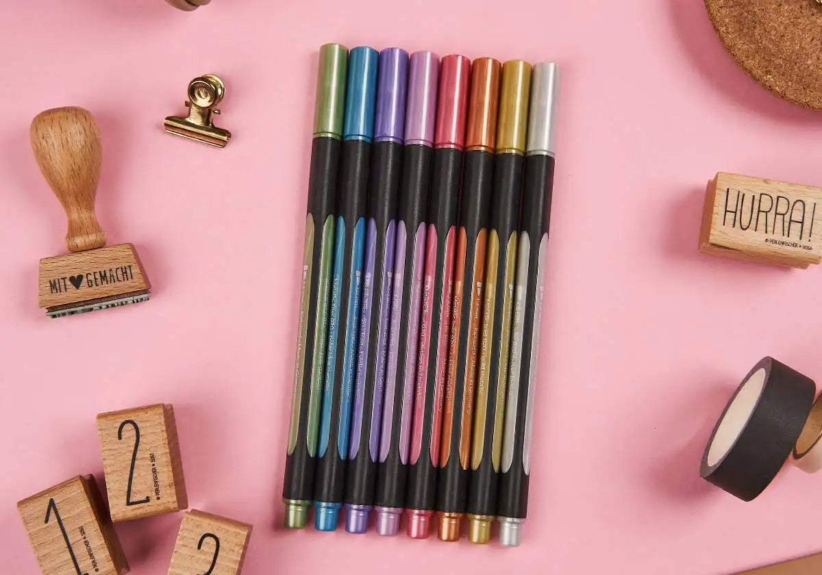 Auf einem rosa Untergrund präsentiert sich das »Nachtglanz« Metallicliner 8er-Set von Schneider. Rund um die farbenfrohen Stifte befinden sich Holzstempel, ein Clip und nummerierte Holzklötze, die der künstlerischen Arbeitsplatzatmosphäre eine kreative Note verleihen.