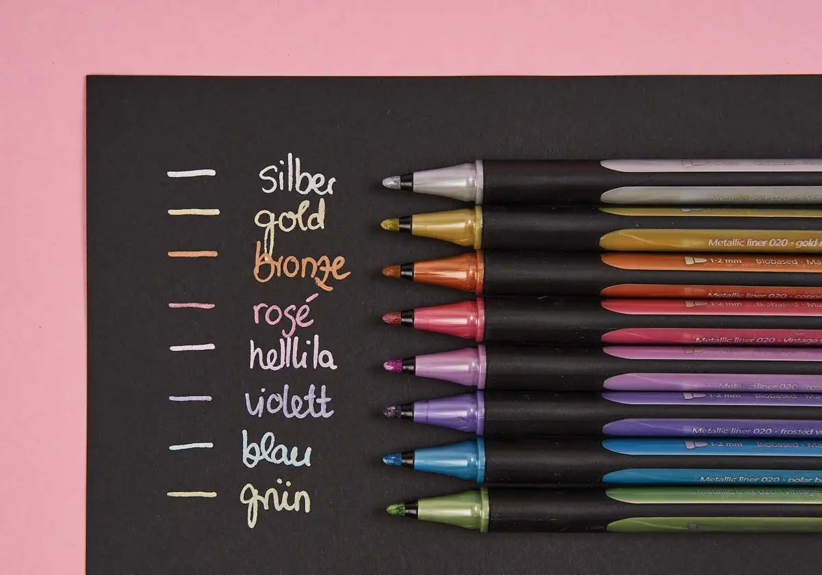 Auf einem schwarzen Blatt dunklem Papier sind die Farbnamen mit dem »Nachtglanz« Metallicliner 8er-Set von Schneider handgeschrieben und verleihen jedem Farbton eine kreative Note: Silber, Gold, Bronze, Roségold, Helllila, Violett, Blau und Grün. Zu jedem Farbton gibt es den passenden Metallicliner auf rosa Untergrund.