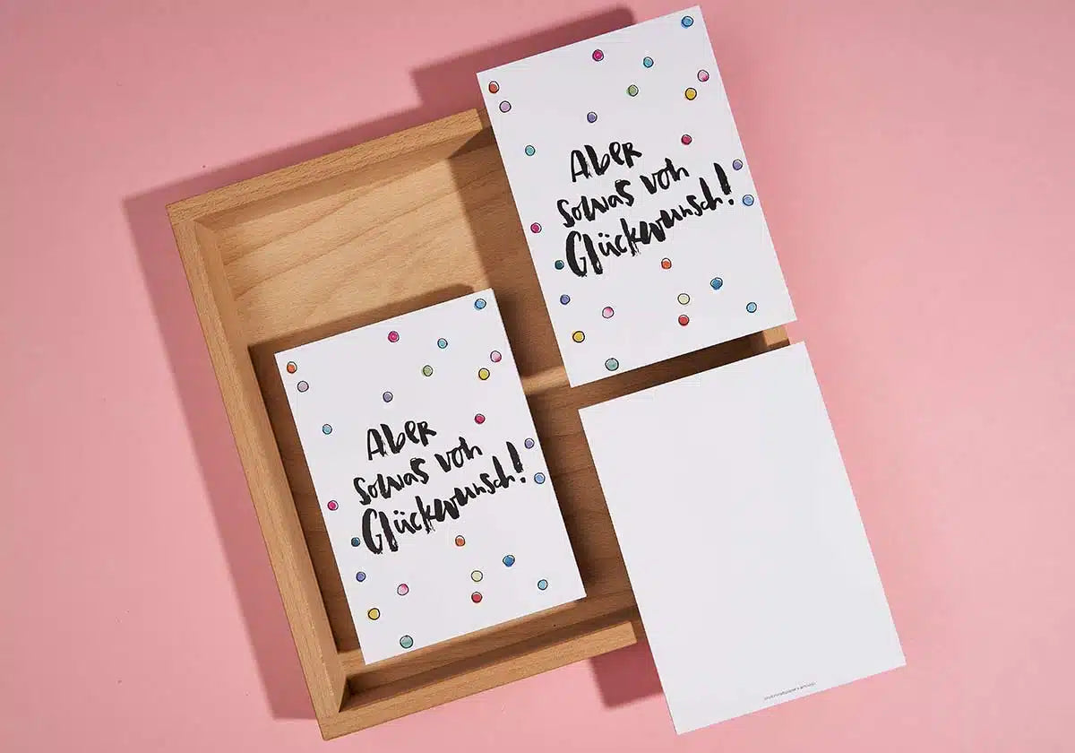 Zwei «Aber sowas von Glückwunsch»-Grußkarten und eine Postkarte von brushmeetspaper x amoedo liegen auf einer rosa Unterlage und einem Holztablett. Jede Karte ist mit bunten Punkten verziert und trägt in fetten schwarzen Lettern den Satz «Aber sowas von Glückwunsch!». Eine leere Karte lugt hervor, bereit, freudige Momente festzuhalten.