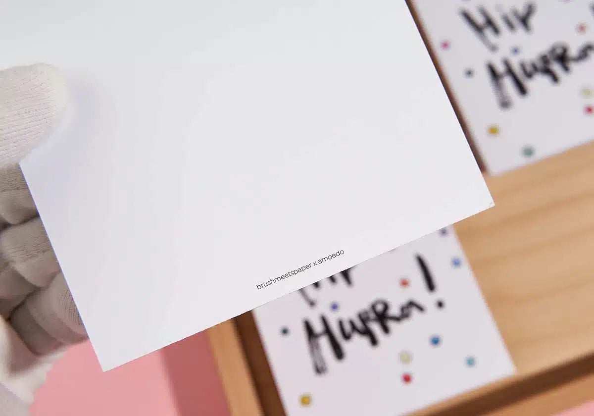 Eine behandschuhte Hand hält eine schlichte weiße Karte mit kleinem Text am unteren Rand, der ein Gefühl von Lebensfreude hervorruft. Der Hintergrund zeigt eine verschwommene Postkarte »Hip Hip Hurra« von brushmeetspaper x amoedo, verziert mit bunten Punkten und teilweise sichtbarem schwarzem Text.