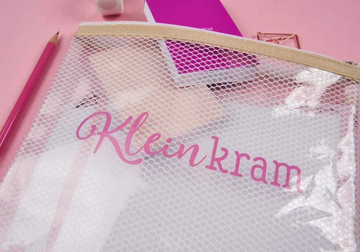 Das Mäppchen »Kleinkram« von amoedo besticht durch seine robuste Oberfläche und den praktischen Reißverschluss. Es ist mit verschiedenen Kleinigkeiten wie Notizbüchern und Schreibwaren gefüllt. Es ist auf einem rosa Untergrund platziert und hat einen rosa Bleistift daneben.