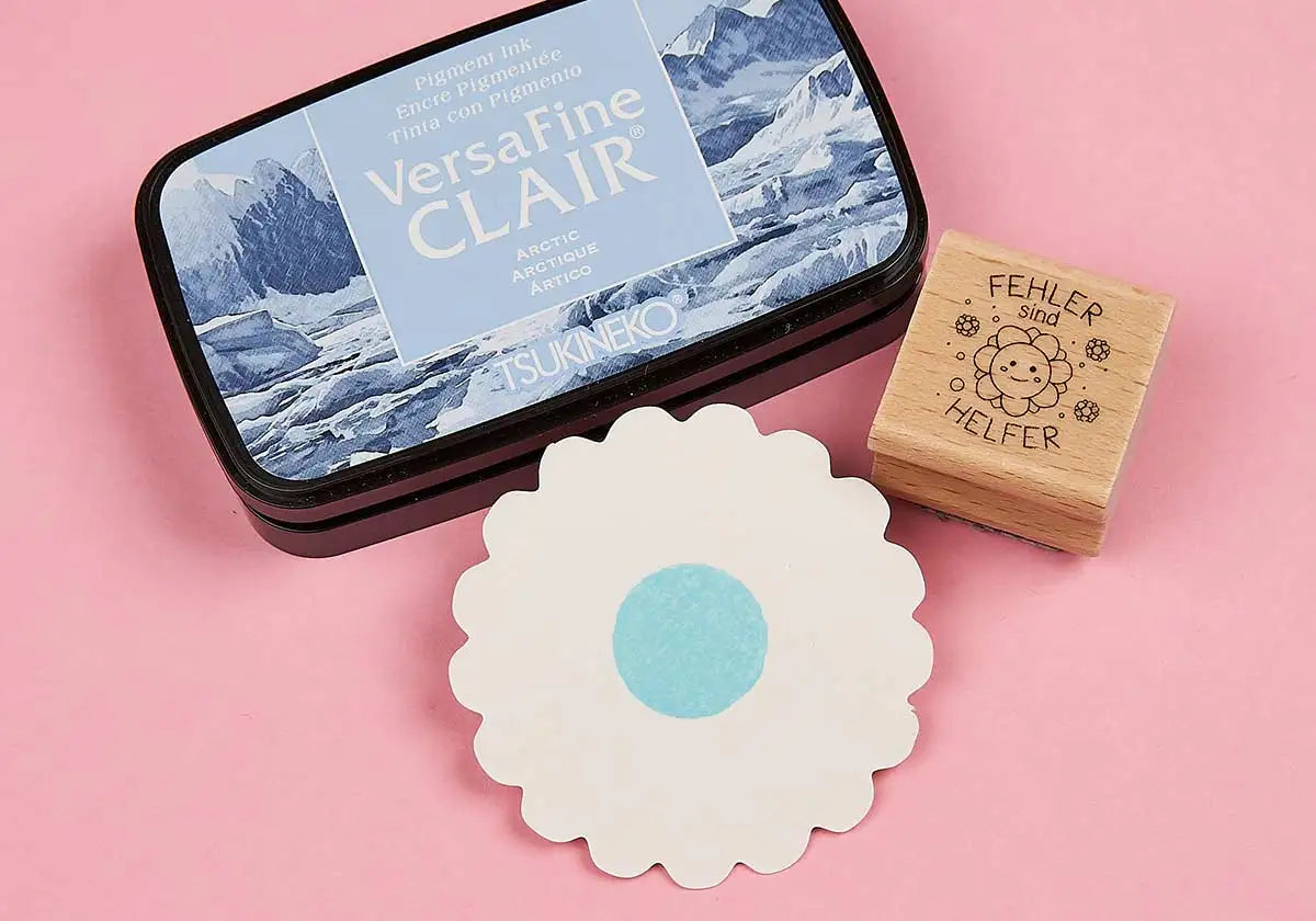 Ein Tsukineko Stempelkissen »VersaFine Clair« in der Farbe Arctic, bekannt für seine stark pigmentierten Stempelabdrücke, wird neben einem Stempel mit einem Smiley und dem deutschen Satz „FEHLER sind HELFER“ sowie einem weißen Papierausschnitt mit einem blauen Kreis in der Mitte, alles angeordnet auf einer rosa Oberfläche, angezeigt.